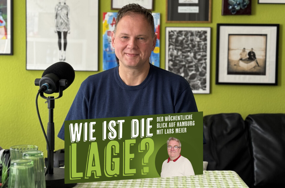 Mann mit Plakat „Wie ist die Lage?“ vor grüner Wand mit Bildern und Mikrofon.
