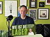 Mann mit Plakat „Wie ist die Lage?“ vor grüner Wand mit Bildern und Mikrofon.