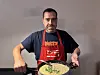 Mann in roter Schürze hält einen Topf mit heller Suppe in der Küche.