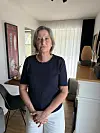 Frau steht in hellem Wohnraum vor Fenster, neben Stuhl, Kommode und Dekoration.