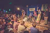 Band spielt auf kleiner Bühne vor sitzendem und stehenden Publikum im Freien.