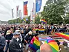 Menschenmenge mit Regenbogenfahnen auf einem Platz vor Fahnenmasten und Bäumen.