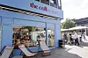 Café „the coffe spot“ mit Außensitzen an einem sonnigen Platz, Passanten gehen vorbei.