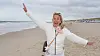 Lachende Frau in weißer Jacke mit ausgebreiteten Armen am Strand vor Meer und Himmel.