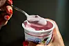 Hand hält Kirschjoghurtbecher, daneben ein Löffel mit cremigem Joghurt.