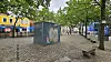 Gepflasterter Platz mit Graffiti-Container, Bäumen und Bänken vor farbigen Gebäuden.