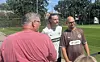 Drei Männer sprechen am Rande eines Fußballtrainingsplatzes, im Hintergrund Rasen, Bäume und Zaun.
