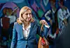 Frau im blauen Blazer steht vor farbiger Wand und hält eine Tasche.