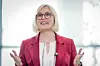 Frau in pinkem Blazer und Brille spricht mit erhobenen Händen vor hellem Hintergrund.