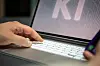 Hand tippt auf Laptop mit KI-Schriftzug, daneben Smartphone im Vordergrund.