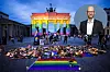 Vor dem Brandenburger Tor liegen Blumen und Kerzen auf einem Regenbogen-Mahnmal; rechts oben ein Porträt eines Mannes.