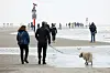 Passanten gehen am Strand von St. Peter-Ording; ein Hund läuft im Vordergrund, im Hintergrund ein Hinweisschild.