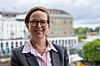 Frau mit Brille und Blazer vor unscharfer Stadtkulisse am Wasser.