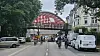 Radfahrer fahren unter einer roten Brücke durch eine Stadtstraße mit geparkten Autos und Häusern.