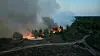 Flammen und Rauch über einem bewaldeten Hang neben einer kurvigen Straße.