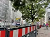 Absperrgitter sichern eine Baustelle an einer Straße mit Bäumen und modernen Gebäuden; Passanten gehen im Hintergrund.