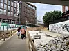 Menschen gehen auf einem Holzsteg an einer Baustelle vorbei, neben Absperrungen und Graffiti.