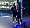 Zwei Sportler sprechen in einer Halle vor einer Sponsorentafel, ein weiterer sitzt im Vordergrund.