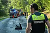 Polizist mit gelber Einsatzweste vor abgesperrter Unfallstelle auf einer Landstraße mit Einsatzfahrzeugen und Trümmern.