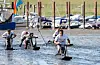 Teilnehmer rutschen mit Holzschlitten durchs Watt in Dangast, Boote liegen im Hintergrund.