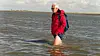 Mann in roter Jacke watet mit Rucksack durch flaches Küstenwasser.