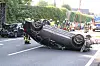 Auf dem Dach liegendes Auto auf einer Straße, Einsatzkräfte sichern die Unfallstelle im Hintergrund.