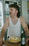 Tätowierter Mann im weißen Tanktop am Tresen mit Pommes, Teller und Bierflasche vor Coca-Cola-Kühlschrank.