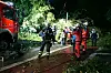 Feuerwehrkräfte stehen nachts auf einer nassen Straße neben einem roten Löschfahrzeug im Wald.