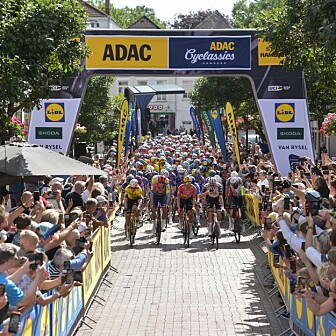 Radrennfeld fährt unter ADAC-Startbogen durch eine von Zuschauern gesäumte Straße.