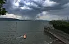 Dramatische Regenwolken über Badegästen am Bodensee und einem Steg am Ufer.