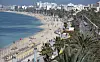 Langer Strand mit Promenade, Sonnenschirmen und Palmen an einer Küstenstadt.