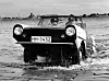 Hamburger Amphicar fährt aus der Elbe ans Ufer, Wasser spritzt, im Hintergrund die Hafenkulisse.
