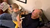 Älterer Mann sitzt auf dem Sofa, Kleinkind steht vor ihm und spielt mit einem Papprollen-Objekt.