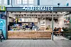 Café „HAFENKATER“ mit offener Theke, Sitzplätzen und Gästen vor einer hellen Fassade.