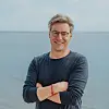 Lächelnder Mann mit Brille steht mit verschränkten Armen am Meer.