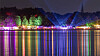 Bunte Lichtprojektionen beleuchten Bäume am Ufer, farbige Spiegelungen auf dem Wasser bei Nacht.