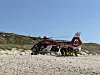 Rettungshubschrauber des ADAC am Strand, Einsatzkräfte arbeiten daneben vor Dünen und blauem Himmel.