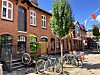 Fahrräder vor roten Backsteinhäusern, darüber bunte Wimpel und ein Baum an sonniger Straße.