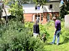 Zwei Personen stehen in einem verwilderten Garten vor einem Wohnhaus und arbeiten zwischen hohem Grün.