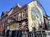Bunte Eckfassade mit großem Wandgemälde, Graffiti, Schildern und mehreren Fenstern an einer Straßenecke.