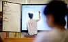 Lehrerin schreibt eine Mathematikaufgabe an der digitalen Tafel, ein Kind ist unscharf im Vordergrund zu sehen.