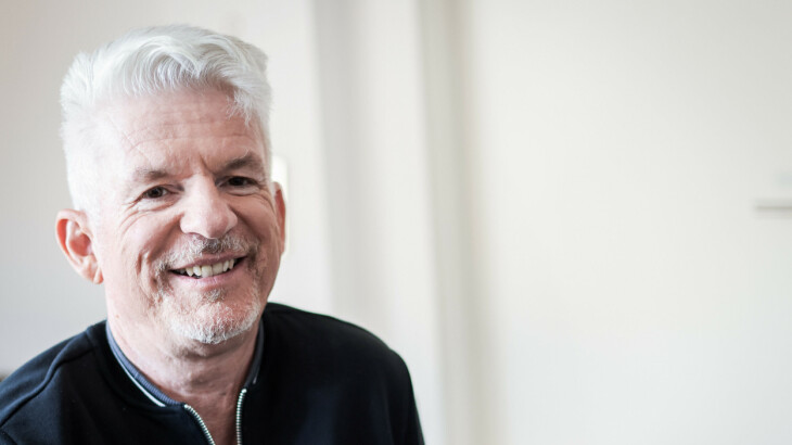 Heinz Strunk lächelt in seiner Wohnung vor hellem Hintergrund.