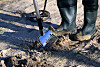 Person sucht mit einem Metalldetektor im Sand; Suchspule und Gummistiefel sind zu sehen.