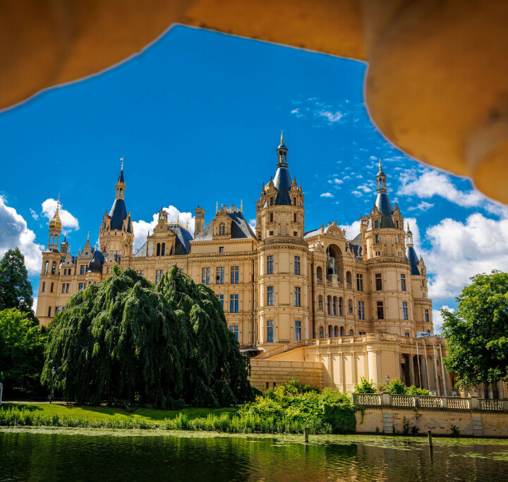 Schweriner Schloss am Burgsee, eingerahmt von einem Steingeländer, bei blauem Himmel und Sonnenschein.