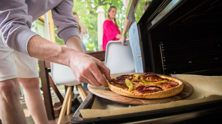 Familie bereitet auf einer Terrasse gemeinsam eine Pizza am Grill zu.
