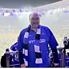 Älterer Fan im blauen Trikot und Schal steht vor einer dicht besetzten Stadiontribüne.