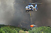 Polizeihubschrauber mit orangefarbenem Löschwasserbehälter über brennendem Wald im Hürtgenwald