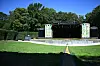 Leere Open-Air-Bühne im Stadtpark, umgeben von Bäumen, Hecken und einer weitläufigen Rasenfläche.