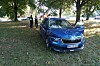 Blauer BMW steht unter Bäumen auf einer Wiese, zwei Personen und Autos sind im Hintergrund zu sehen.