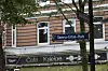 Man sieht ein blaues Straßenschild mit der Aufschrift „Semra-Ertan-Platz“, dahinter ein Wohnhaus, im Erdgeschoss das Café Miller.